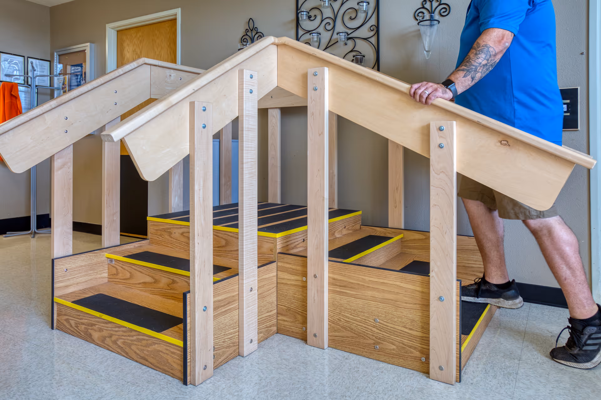 A person in a blue shirt and shorts is using a wooden physical therapy stair stepper with handrails on both sides inside a facility. The stair stepper has three steps with black anti-slip strips and yellow edges. The background shows a beige wall with decorative wall sconces and a door.