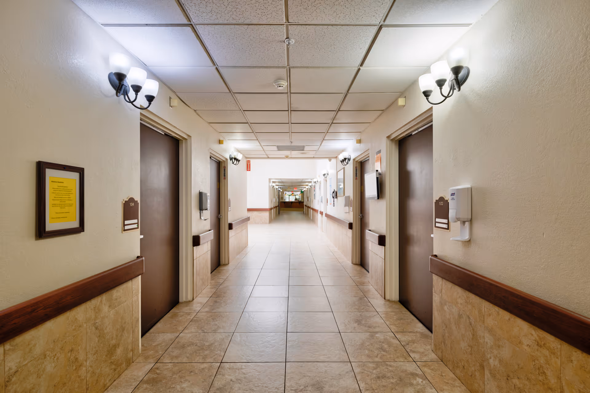 Well-lit tiled hallway with closed doors on both sides, wall sconces, and handrails in a senior living facility.