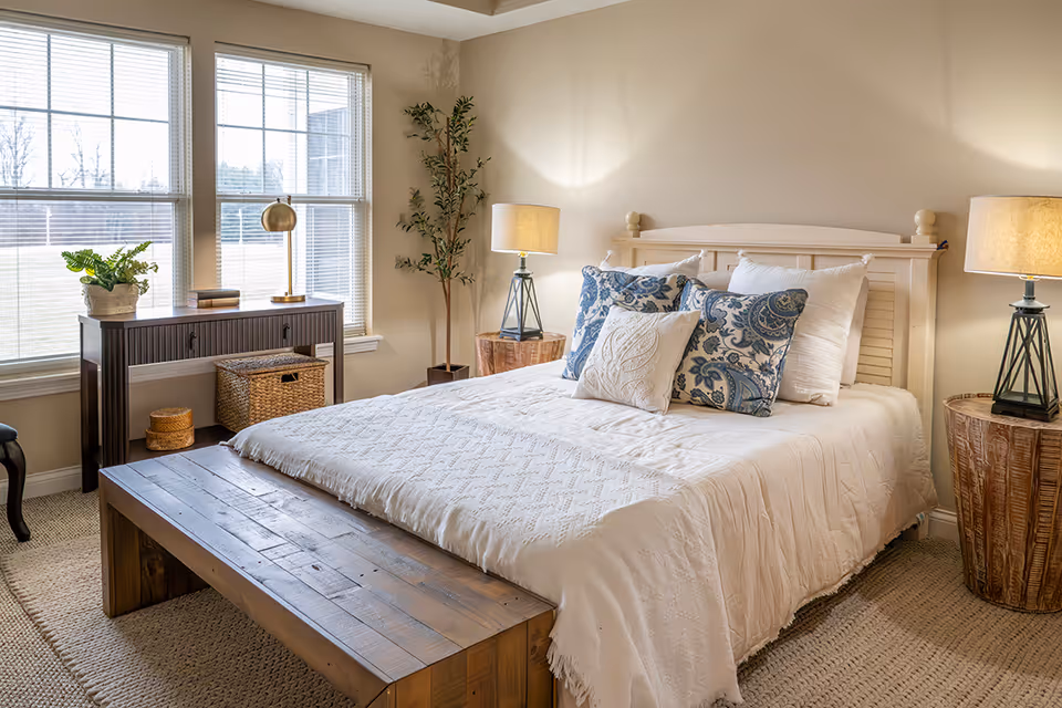 Sunlit bedroom with a white bed topped with patterned pillows, a wooden bench at the foot, bedside lamps, and large windows.