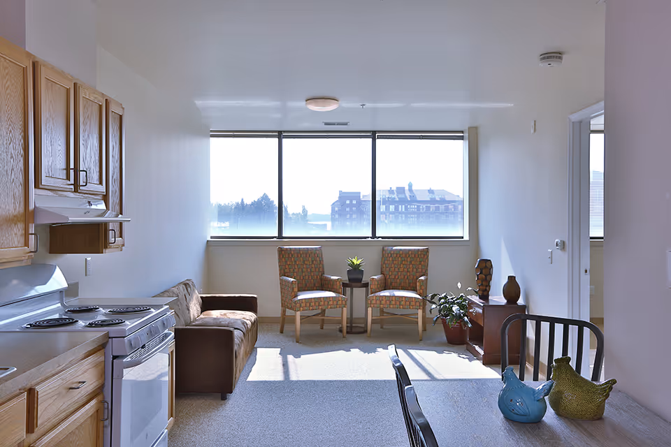 A compact apartment-style living area with a small kitchen on the left, a sofa, two patterned chairs by a large window, and potted plants.