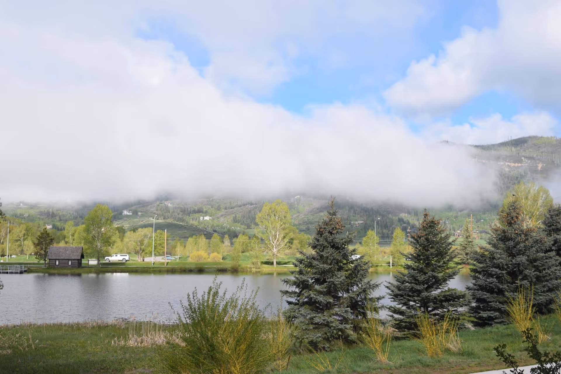 A scenic outdoor view featuring a calm pond with green grass and bushes in the foreground, several evergreen trees, a small wooden cabin, and a white vehicle parked near the water. In the background, there are green hills partially covered by low-hanging clouds under a blue sky with scattered clouds.