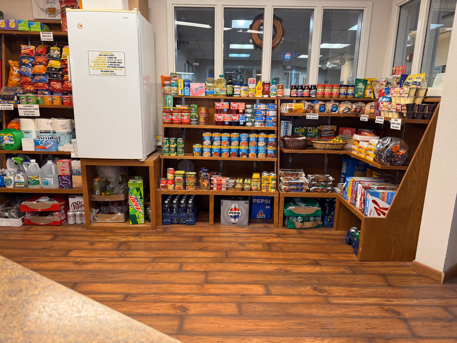 Interior view of a small convenience store or snack shop area with wooden shelves stocked with various canned goods, snacks, beverages, and household items. A white refrigerator is positioned on the left side, and the floor has a wood-like finish. The shelves contain items such as canned Spam, chips, bottled water, soda, laundry detergent, and snack bars.