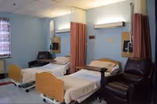A patient room with two hospital-style beds, reclining chairs, and privacy curtains between them.