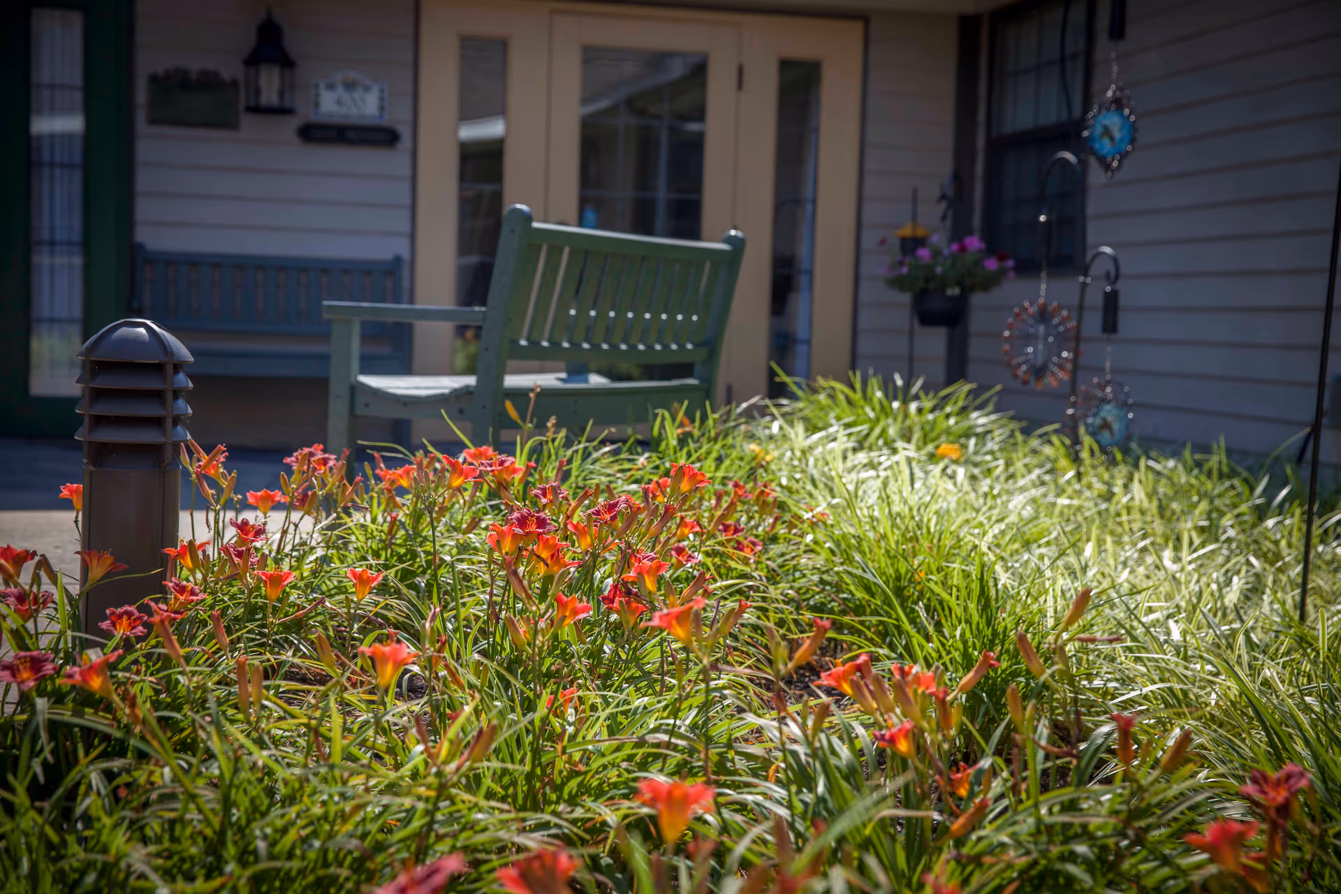 Red-orange daylilies and green foliage in a garden bed in front of a porch with a green bench at a senior living building entrance.