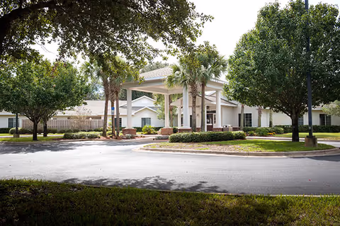Exterior view of a single-story senior living facility building with a covered entrance supported by white columns, surrounded by palm trees and other greenery under a partly cloudy sky.