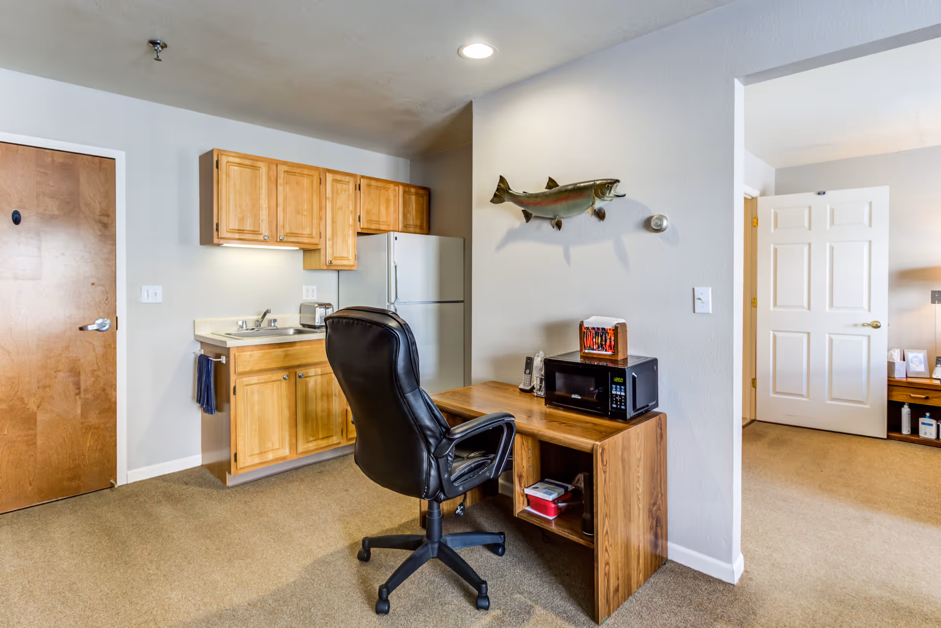 Interior view of a room in Keystone Bluffs Assisted Living featuring a small kitchenette with wooden cabinets, a white refrigerator, and a sink. In front of the kitchenette is a wooden desk with a black office chair, a microwave, and a telephone. A mounted fish decoration is on the wall above the desk. An open doorway leads to another room with a white door and a wooden side table with a lamp and framed photo.