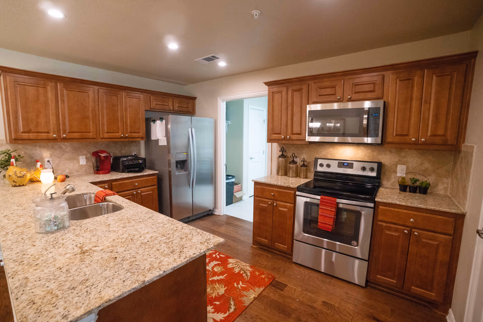 A modern kitchen with wooden cabinets, granite countertops, a stainless steel refrigerator, oven, and microwave. The kitchen features a double sink, a coffee maker, a toaster, and decorative items on the counters. The floor is wooden, and there is a red patterned rug in front of the oven. A doorway leads to another room in the background.