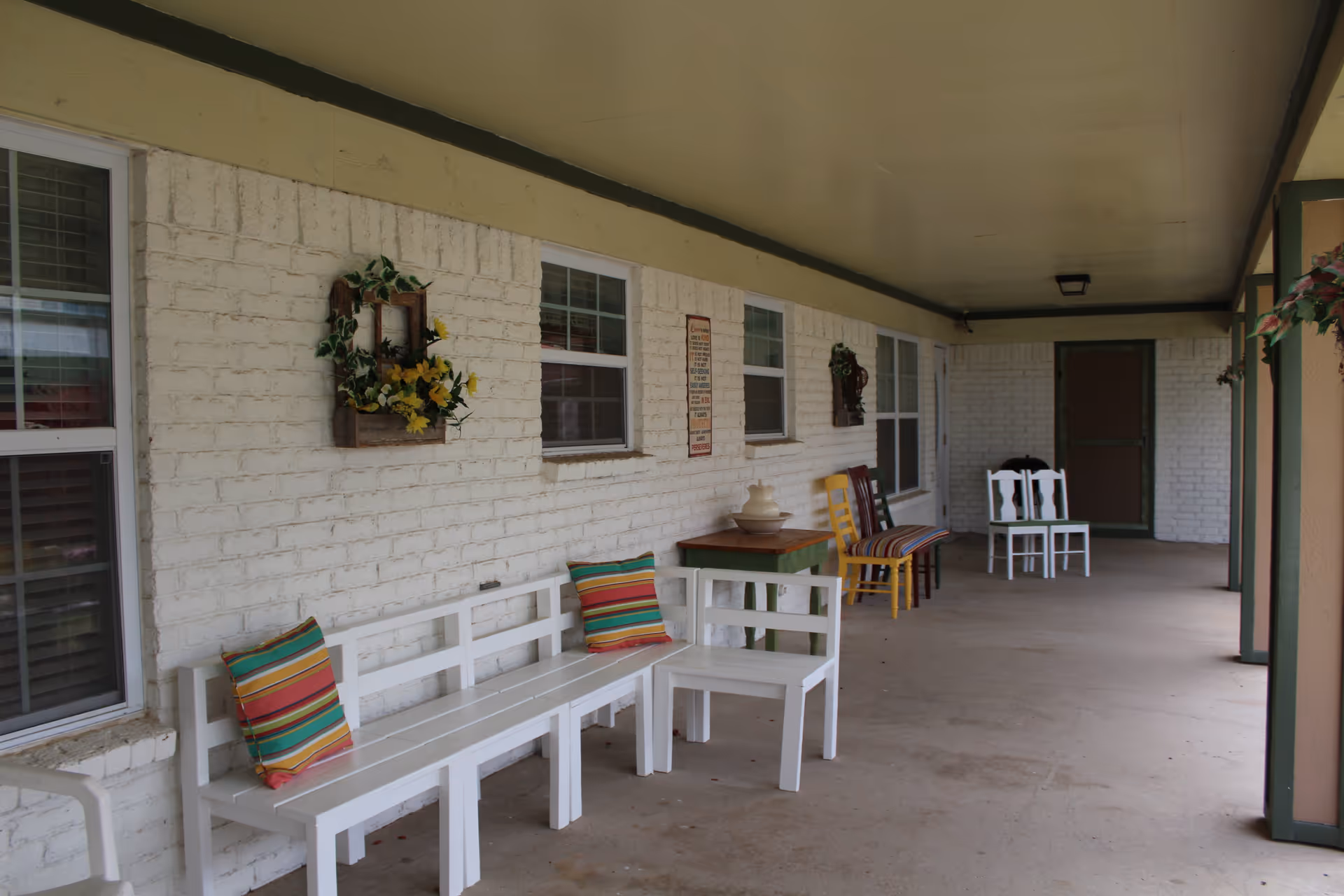 Covered exterior porch with white benches, colorful pillows and chairs arranged along a brick wall under a ceilinged walkway.