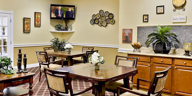 A cozy dining area with wooden tables and chairs arranged on a patterned carpet. The walls are decorated with framed pictures, a decorative plate arrangement, and a small mounted TV. There are plants and floral centerpieces on the tables and countertop, creating a welcoming atmosphere.