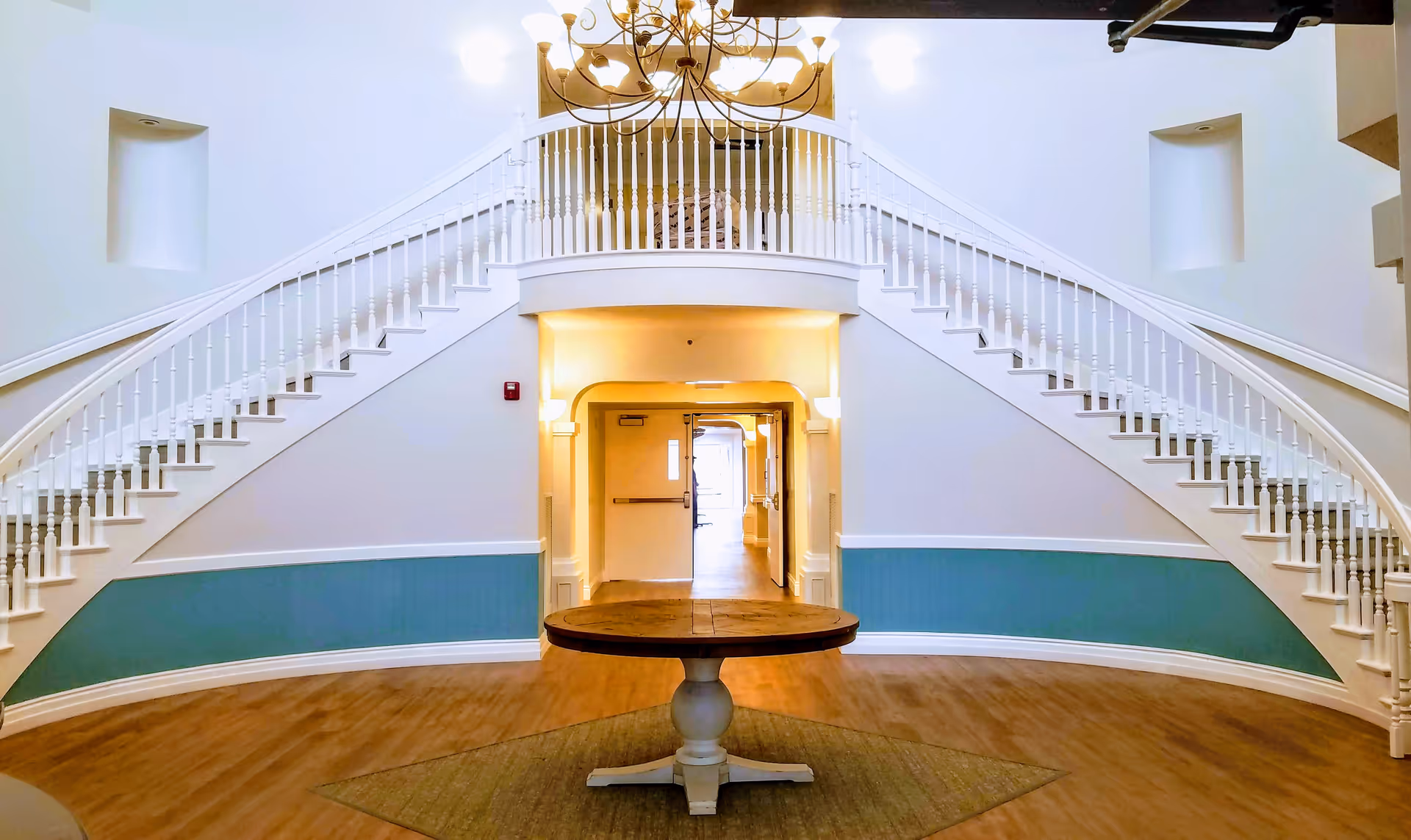 Interior view of a senior living facility lobby with a central round wooden table on a rug, flanked by two symmetrical white staircases with railings leading to an upper balcony. The walls are painted white with a teal accent panel, and a chandelier hangs from the ceiling.