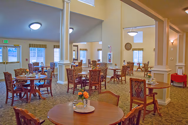 A spacious dining room with multiple round wooden tables and ornate wooden chairs. Each table has a small floral centerpiece and table number. The room has beige walls, carpeted floors, and several windows allowing natural light to enter. Ceiling lights provide additional illumination.
