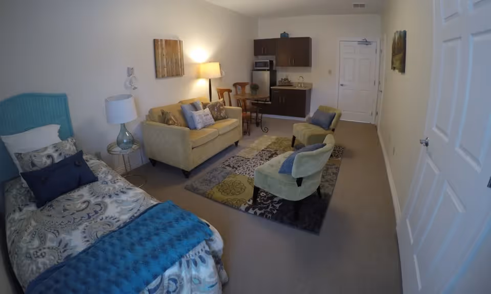 Studio-style senior living apartment showing a bed on the left, a sofa and two chairs around a rug, and a small kitchenette at the back.