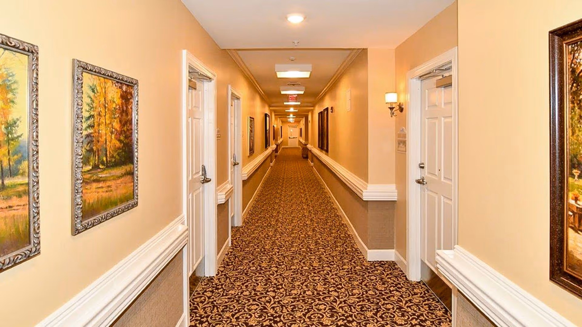 A long, well-lit hallway in a senior living facility with patterned carpet, beige walls, white doors on both sides, and framed paintings hanging on the walls.