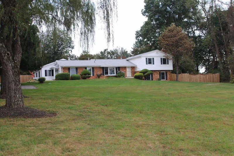 A single-story residential building with a brick and white siding exterior, surrounded by a large grassy lawn and trees. There is a wooden fence on the right side and trimmed bushes near the building.