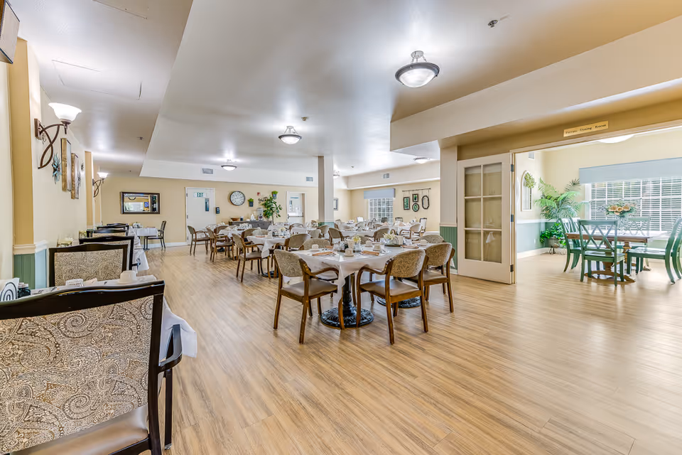 A spacious dining room in a senior living facility with multiple tables covered with white tablecloths and set for meals. The room has wooden flooring, beige walls, and soft lighting from ceiling fixtures and wall sconces. There is an adjoining room visible through open double doors, featuring more dining tables and large windows with blinds, allowing natural light to fill the space. The overall atmosphere is clean, bright, and welcoming.