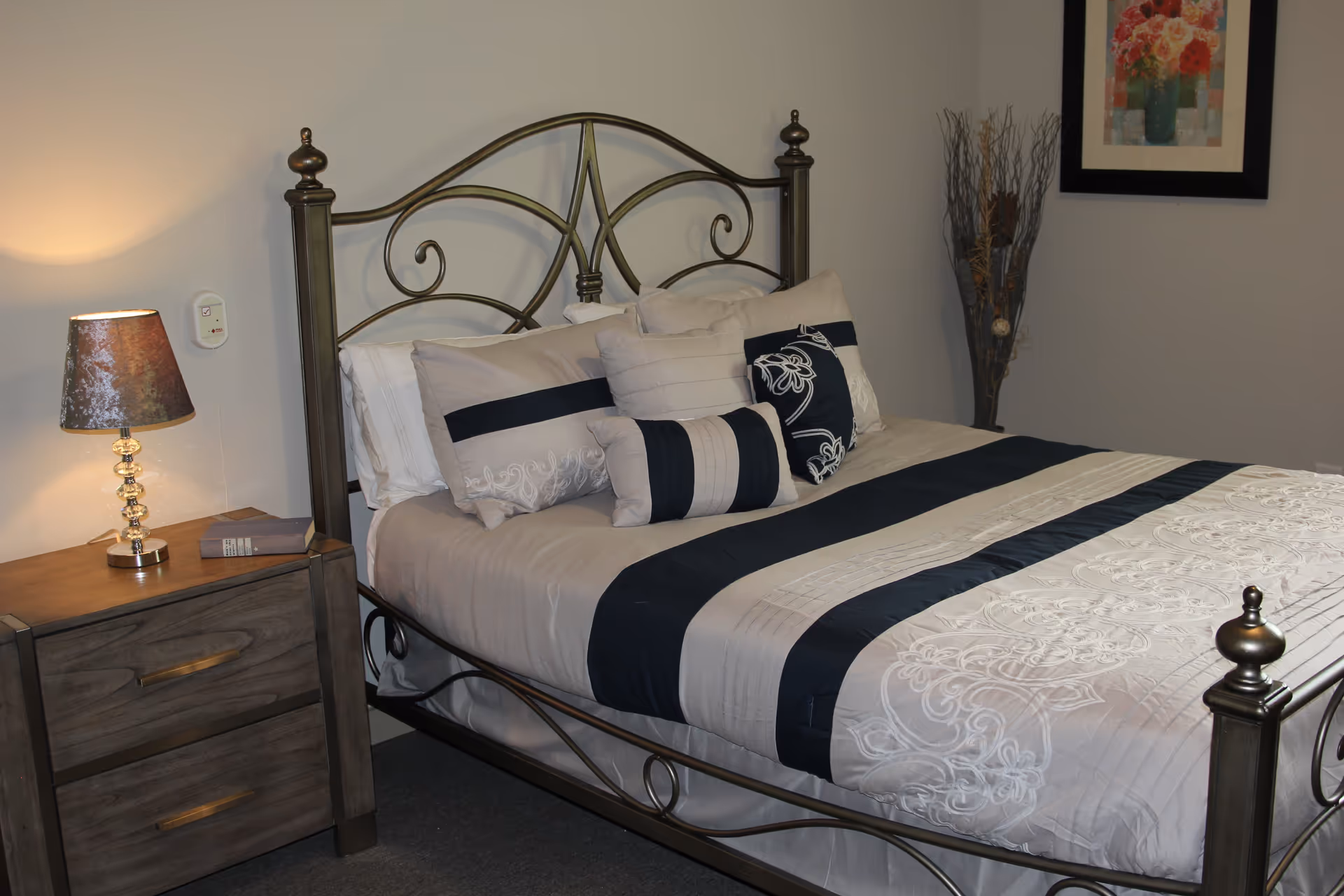 A neatly made bed with a decorative metal headboard and multiple pillows in shades of beige and navy blue. Next to the bed is a wooden nightstand with two drawers, a lamp with a textured brown shade, and a small book. On the wall above the nightstand is a small device, possibly a medical alert button. A framed floral painting and a tall decorative vase with dried branches are visible on the wall behind the bed.