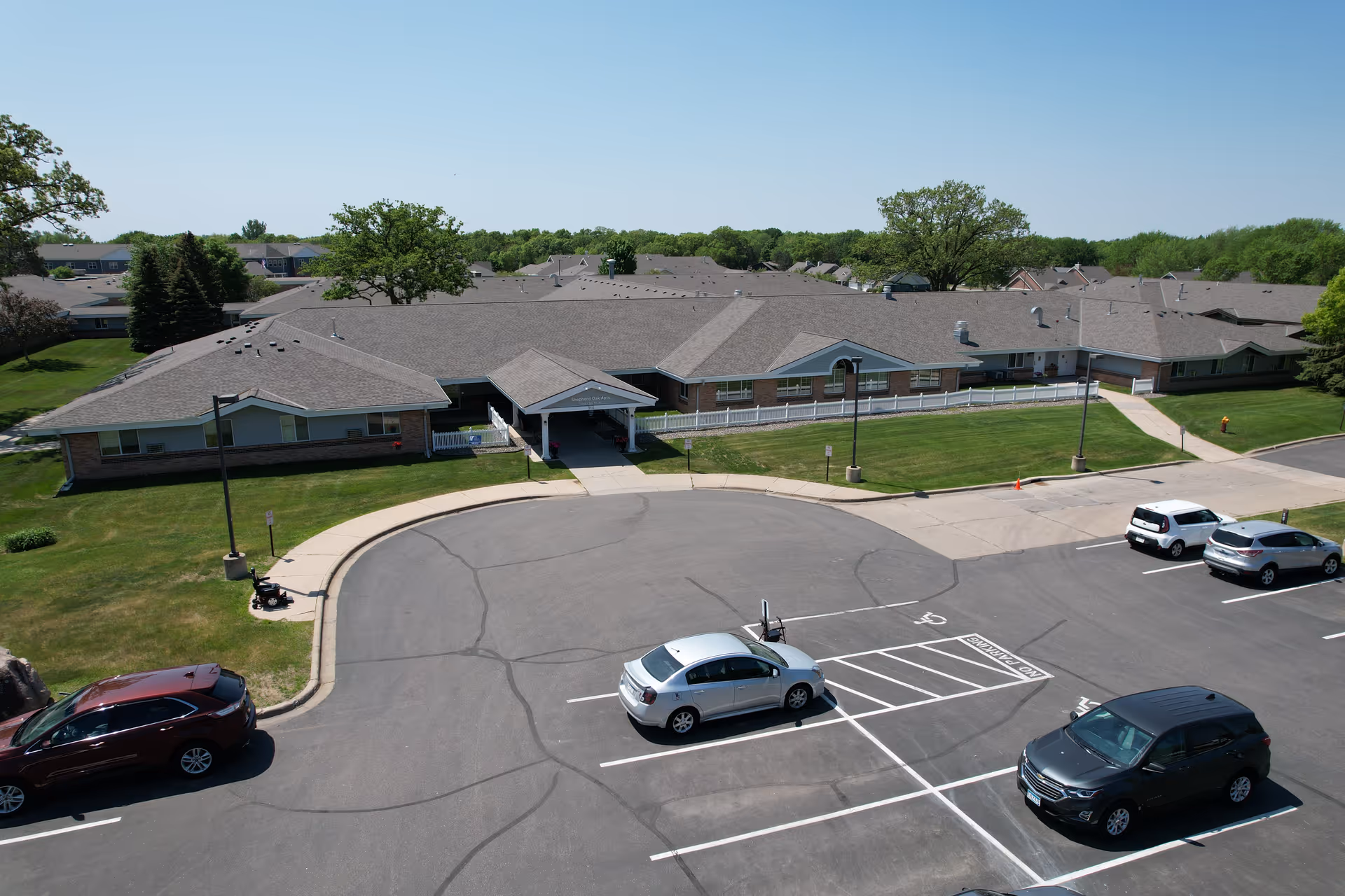 Aerial view of a single-story senior living facility building with a gray roof and brick exterior, surrounded by green lawns and trees. Several cars are parked in the parking lot in front of the building under a clear blue sky.