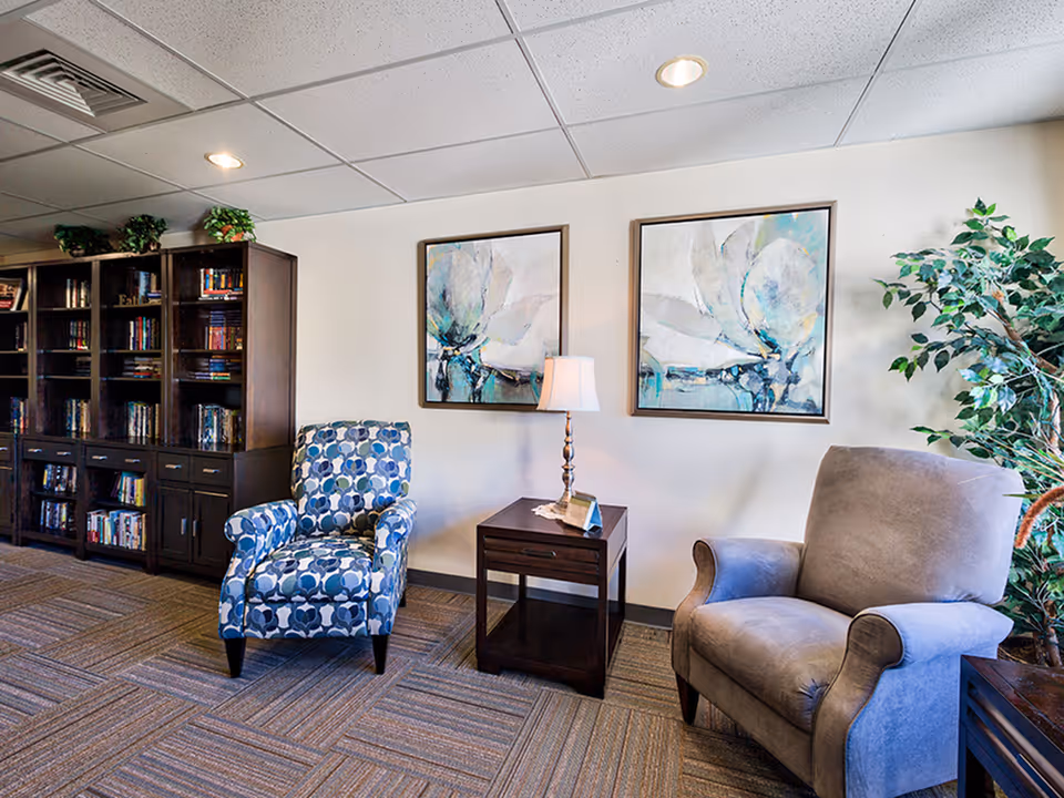 A cozy seating area in a senior living facility with two armchairs, one patterned in blue and white and the other solid gray, separated by a wooden side table with a lamp. Behind the chairs are two framed abstract floral paintings on a white wall. To the left is a dark wooden bookshelf filled with books, and to the right is a tall green potted plant. The floor is carpeted with a patterned design, and the ceiling has recessed lighting.