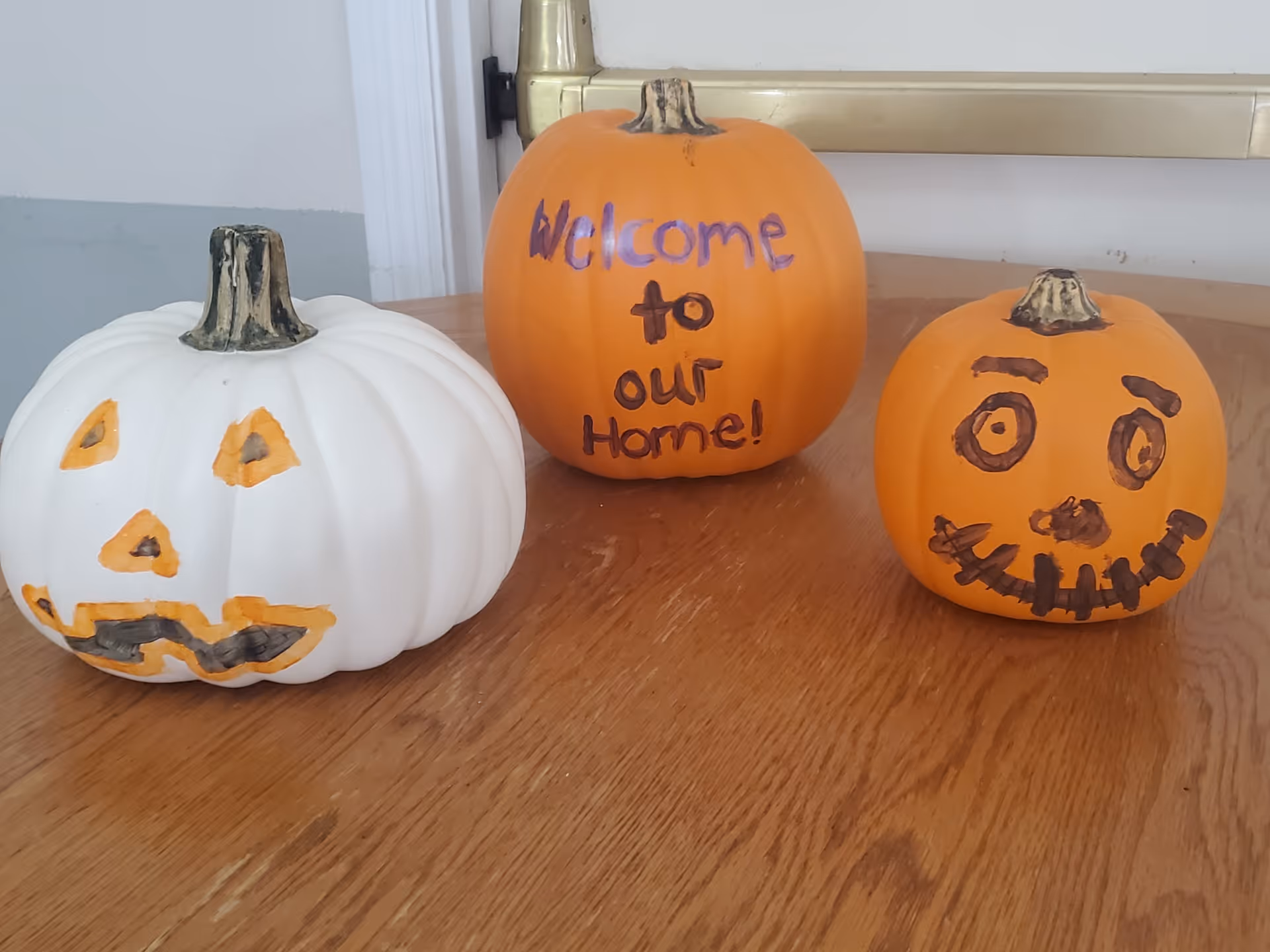 Three small pumpkins on a wooden table. The white pumpkin has a painted jack-o'-lantern face, the middle orange pumpkin has the words 'Welcome to our Home!' written on it, and the right orange pumpkin has a painted face with eyes, eyebrows, nose, and a stitched mouth.