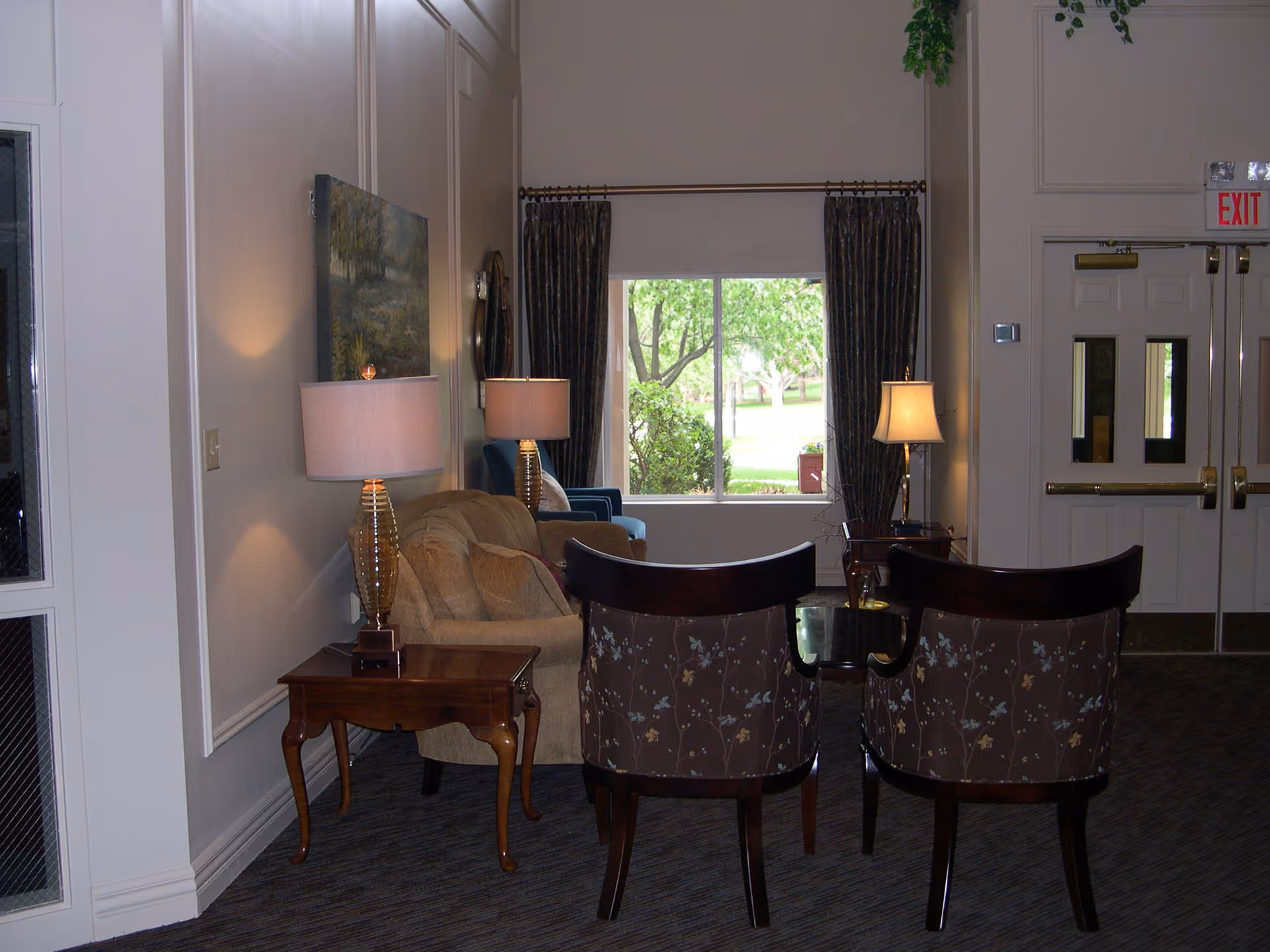 A cozy seating area in a senior living facility with two floral-patterned chairs facing a small table, a beige sofa with cushions, two table lamps with beige shades, a large window with dark curtains showing greenery outside, and double exit doors to the right.