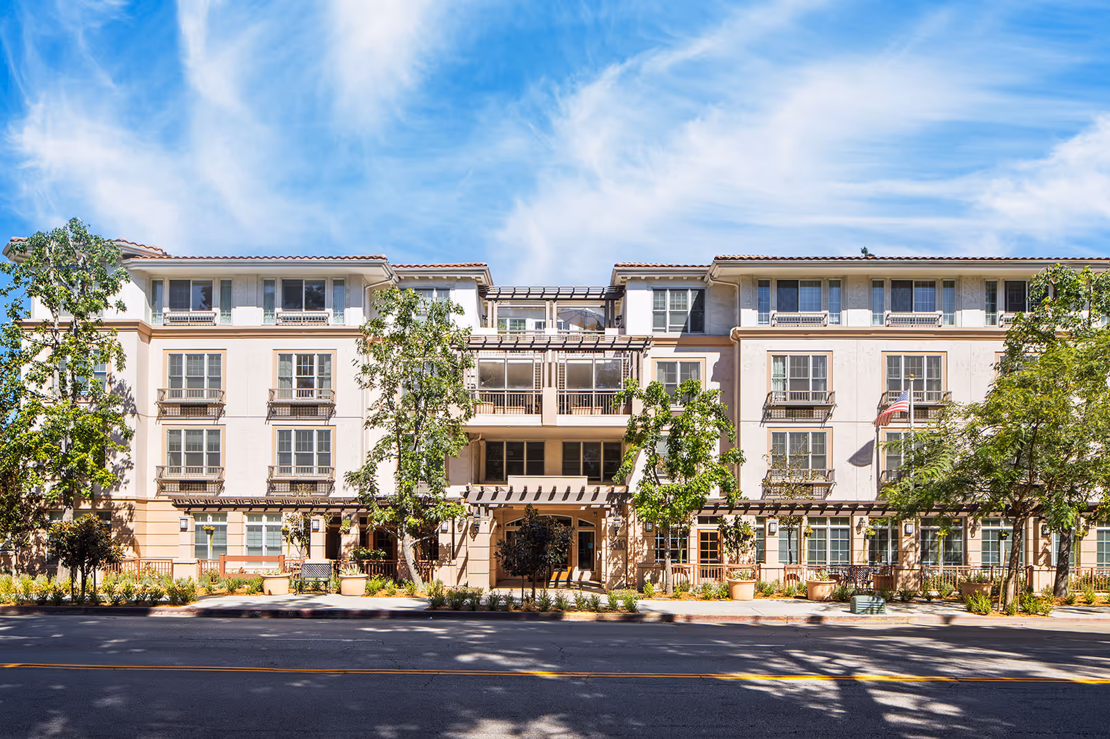 Front exterior of a four-story beige residential building with balconies, trees, and a clear blue sky.