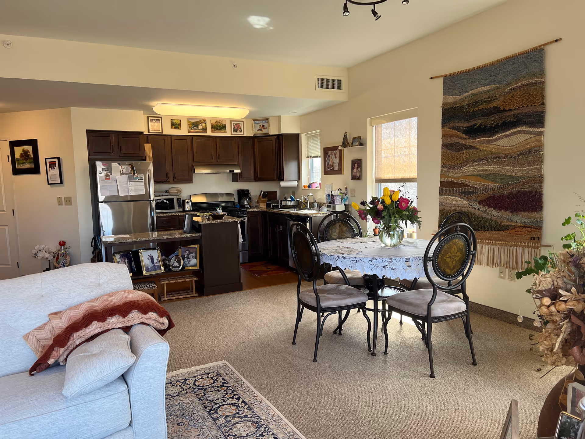 A cozy living space featuring a small kitchen with dark wooden cabinets, stainless steel appliances, and a countertop with framed photos. Adjacent to the kitchen is a round dining table covered with a lace tablecloth and a vase of colorful flowers, surrounded by four chairs. A light-colored sofa with folded blankets is partially visible in the foreground. The room has beige walls, a woven wall hanging, and windows letting in natural light.