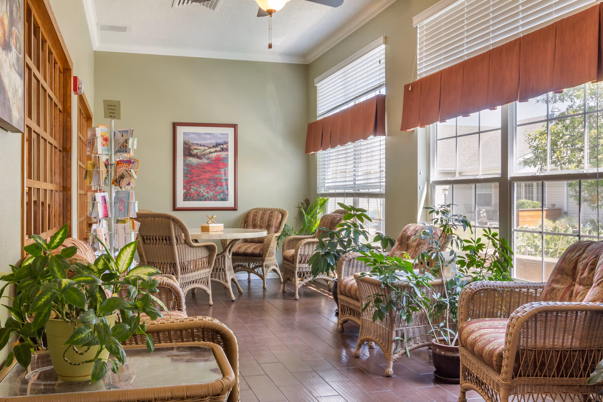 Sunlit communal sitting area with wicker chairs, small tables, potted plants, and large windows.
