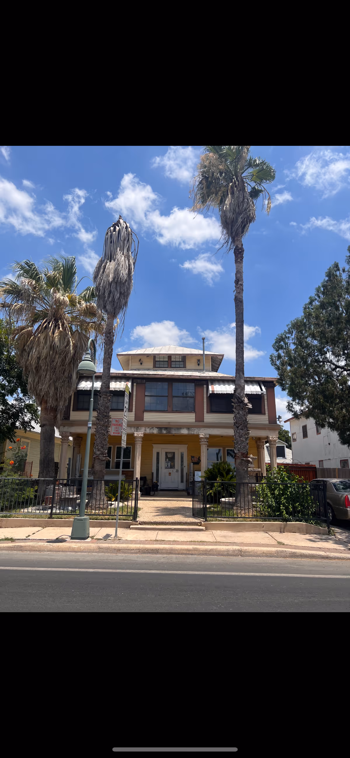Front exterior view of a two-story assisted living facility with a porch, surrounded by palm trees and a small fenced yard under a blue sky with scattered clouds.