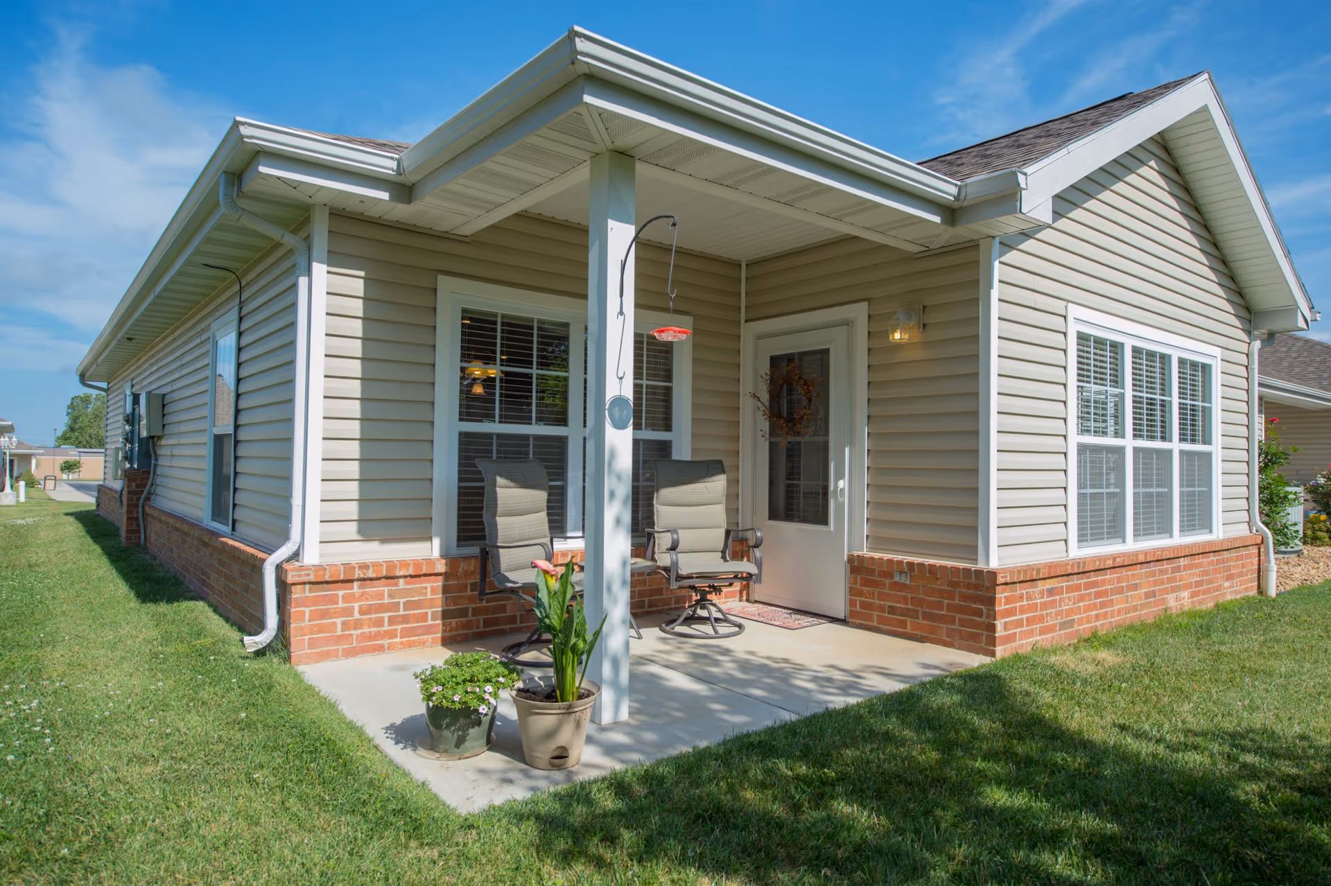 Front porch of a single-story unit with a covered entry, two chairs, potted plants, beige siding and brick foundation.
