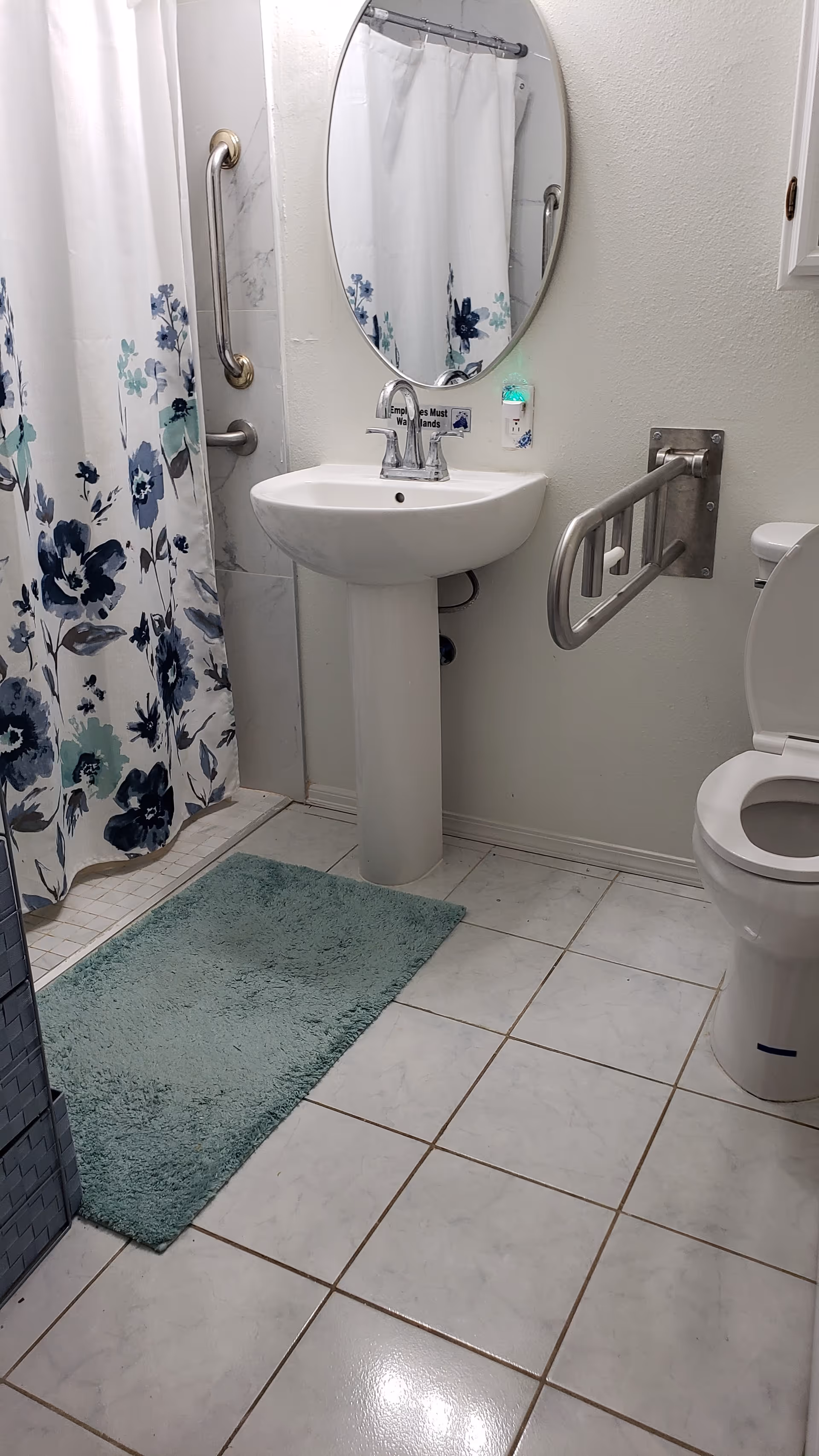 A clean bathroom with a white pedestal sink, an oval mirror above it, a toilet with a safety grab bar on the wall, a shower with a floral curtain in shades of blue and green, and a light green bath mat on the tiled floor.