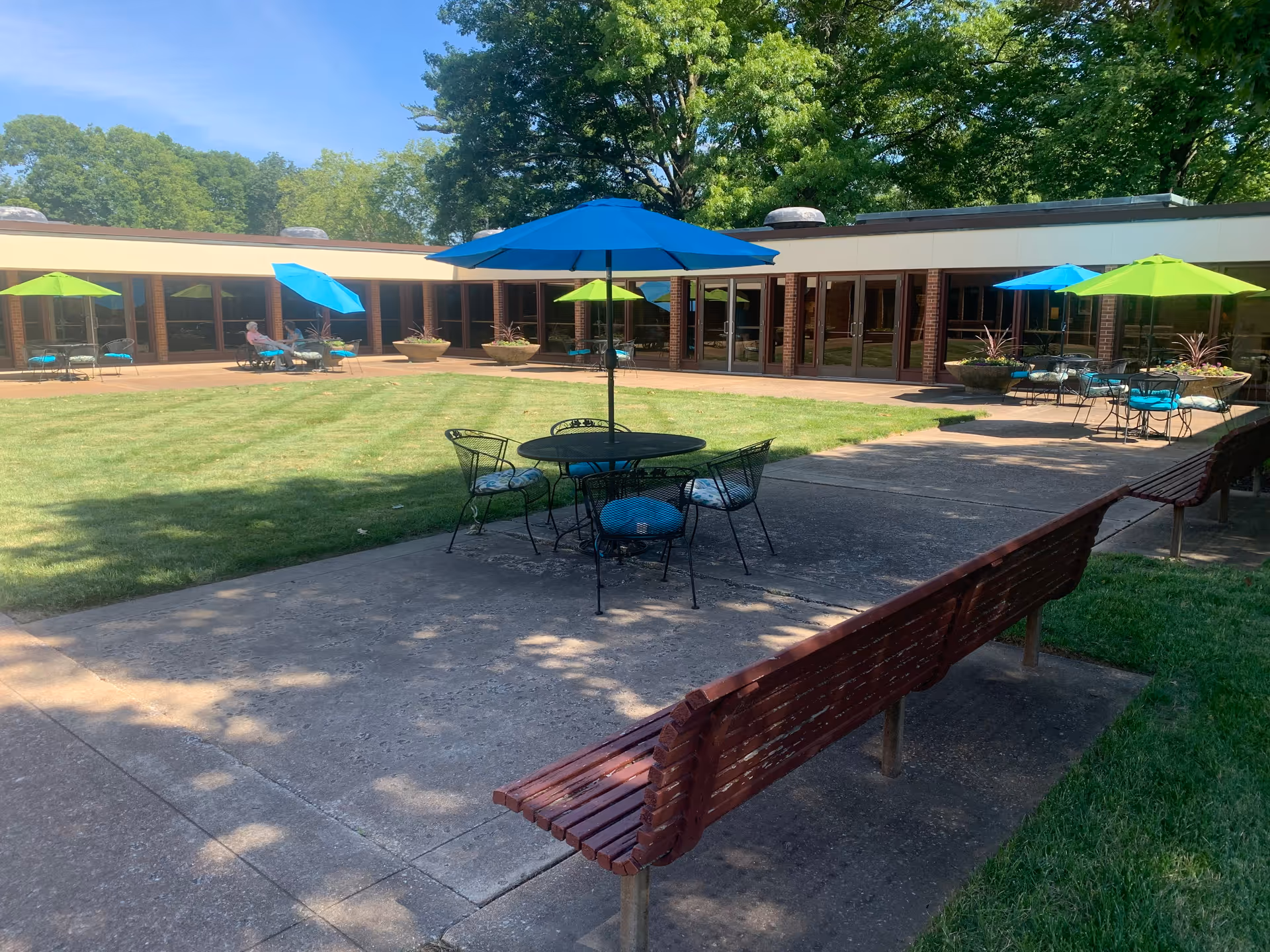 Sunny courtyard with patio tables and colorful umbrellas, benches, and a grassy lawn in front of a low building.