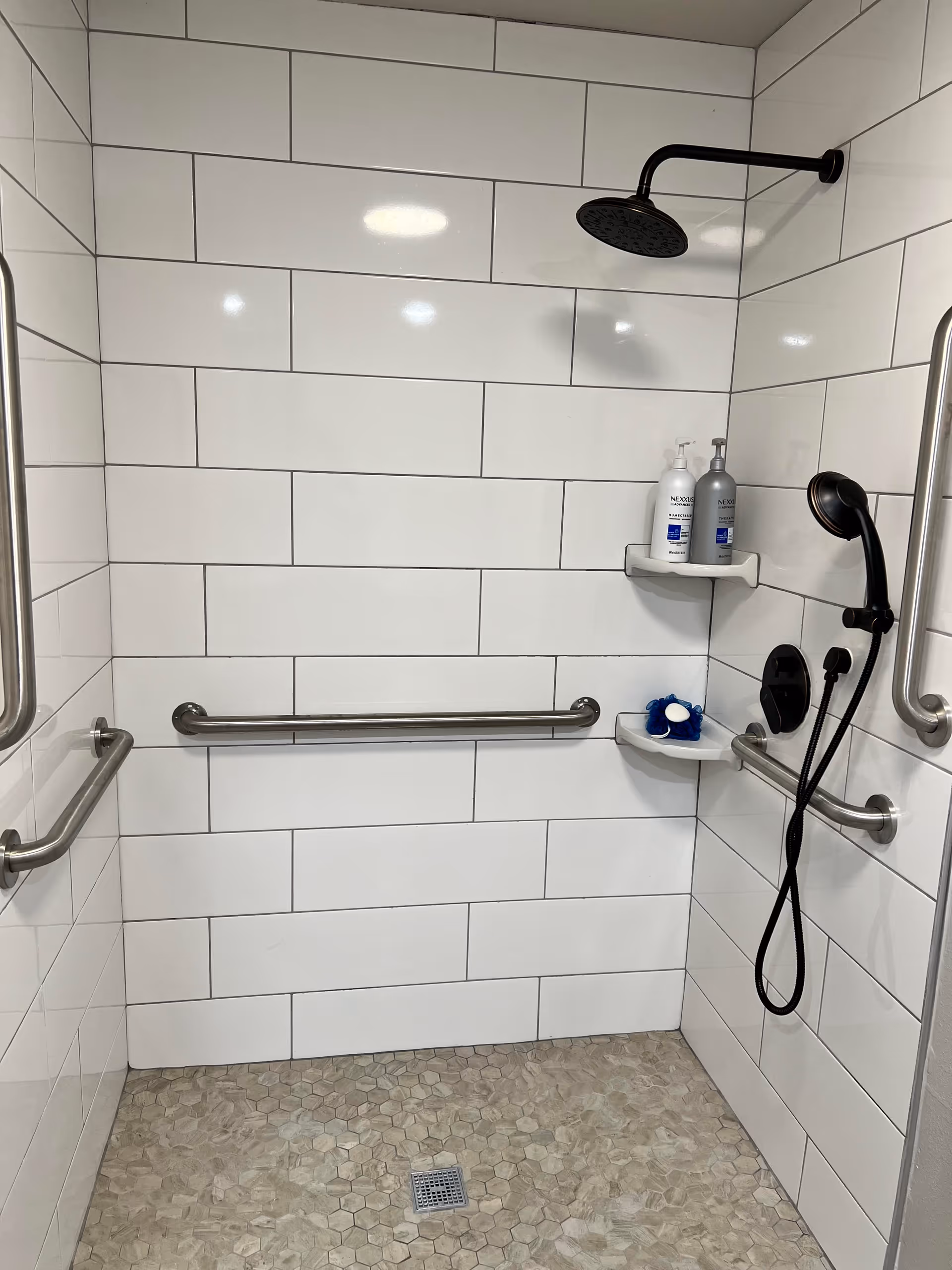 Accessible tiled shower stall with stainless grab bars, overhead rain showerhead, handheld shower, and corner shelves with toiletries.