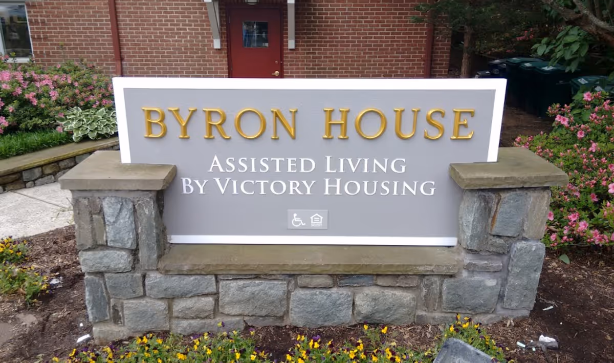 Front sign reads Byron House Assisted Living, set against a brick building with landscaping.