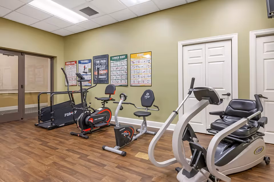 A small exercise room with three pieces of fitness equipment including a treadmill, a stationary bike, and a seated elliptical machine. The room has wood flooring, light green walls, and several exercise instruction posters on the wall. There are two white double doors on the right side and a glass door on the left side.
