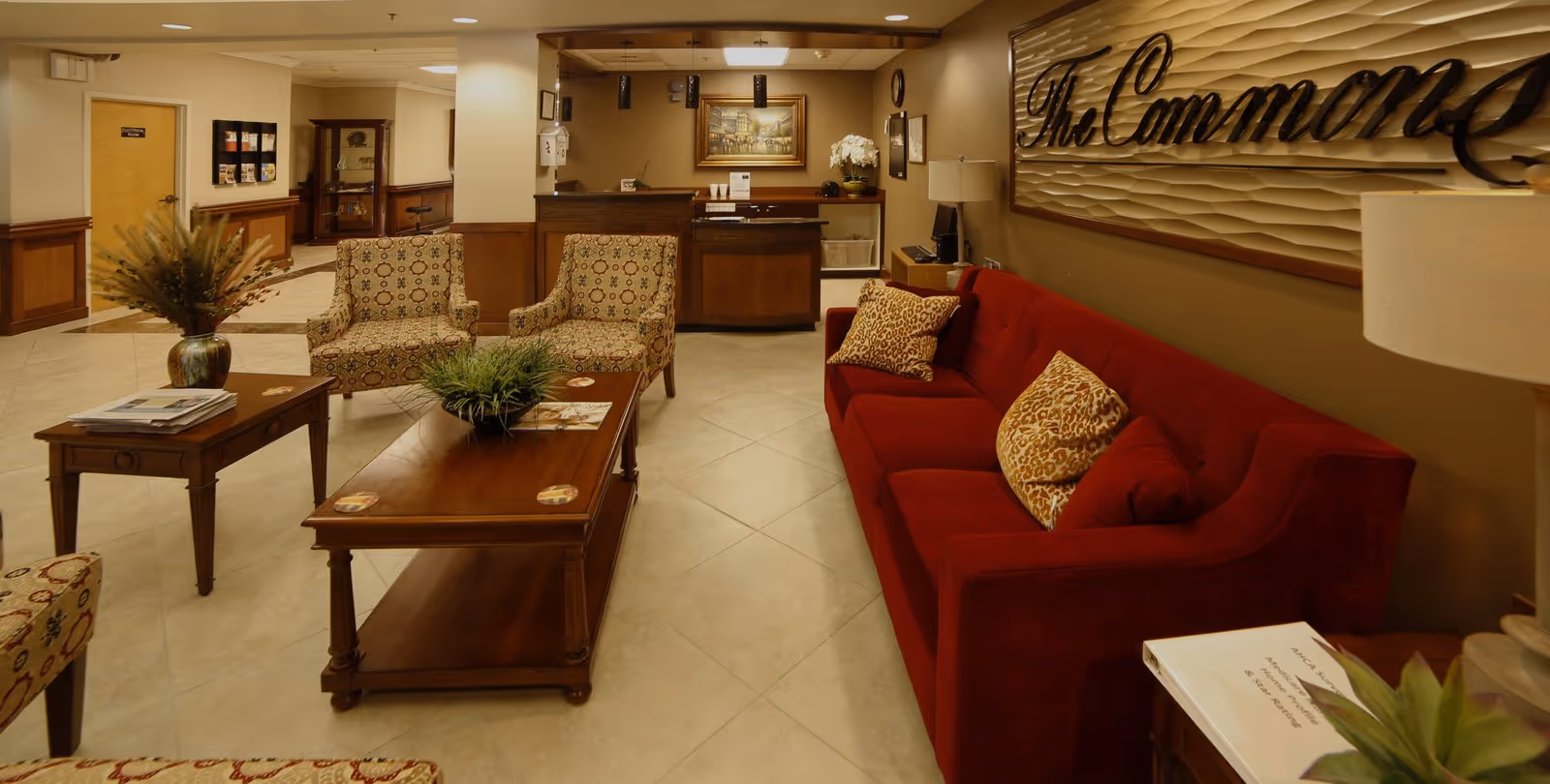 A cozy common area with a red sofa adorned with patterned pillows, two patterned armchairs, and wooden coffee tables with plants and magazines. In the background, there is a reception desk with a painting above it and a sign on the wall that reads 'The Commons'. The room has warm lighting and beige tiled floors.