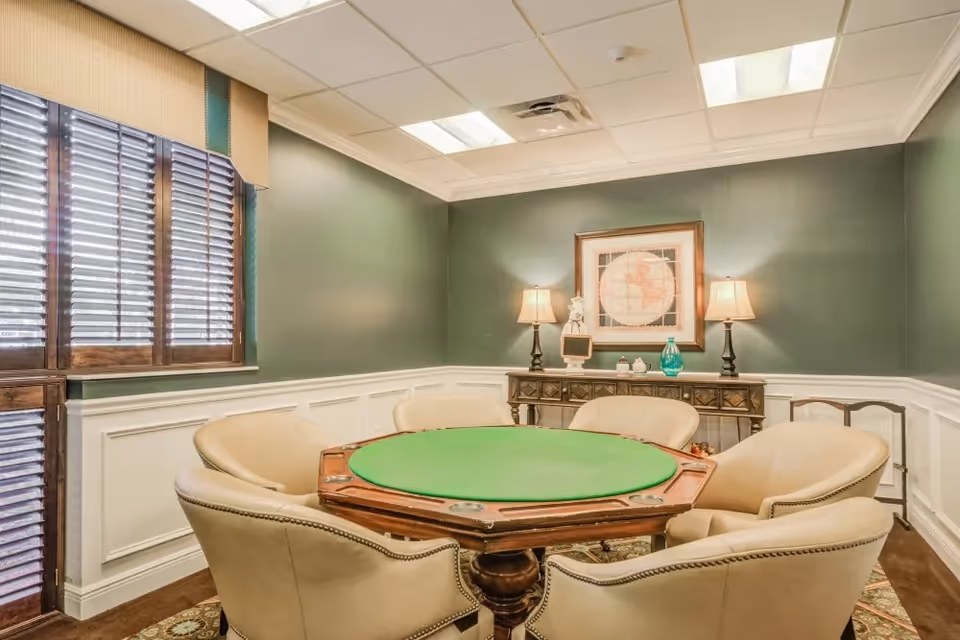A cozy room with a green felt poker table surrounded by six beige upholstered chairs. The walls are painted green with white wainscoting, and there is a wooden sideboard against the back wall with two table lamps, decorative items, and a framed map hanging above it. The room has a window with wooden blinds and a patterned carpet.