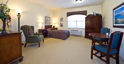 A spacious senior living bedroom with a single bed covered in a patterned bedspread, a green upholstered armchair, a wooden dresser with a lamp and flowers, a round wooden table with two blue cushioned chairs, a large window with a valance, and a wooden armoire. The room is well-lit with ceiling and floor lamps, and decorated with framed artwork on the walls.