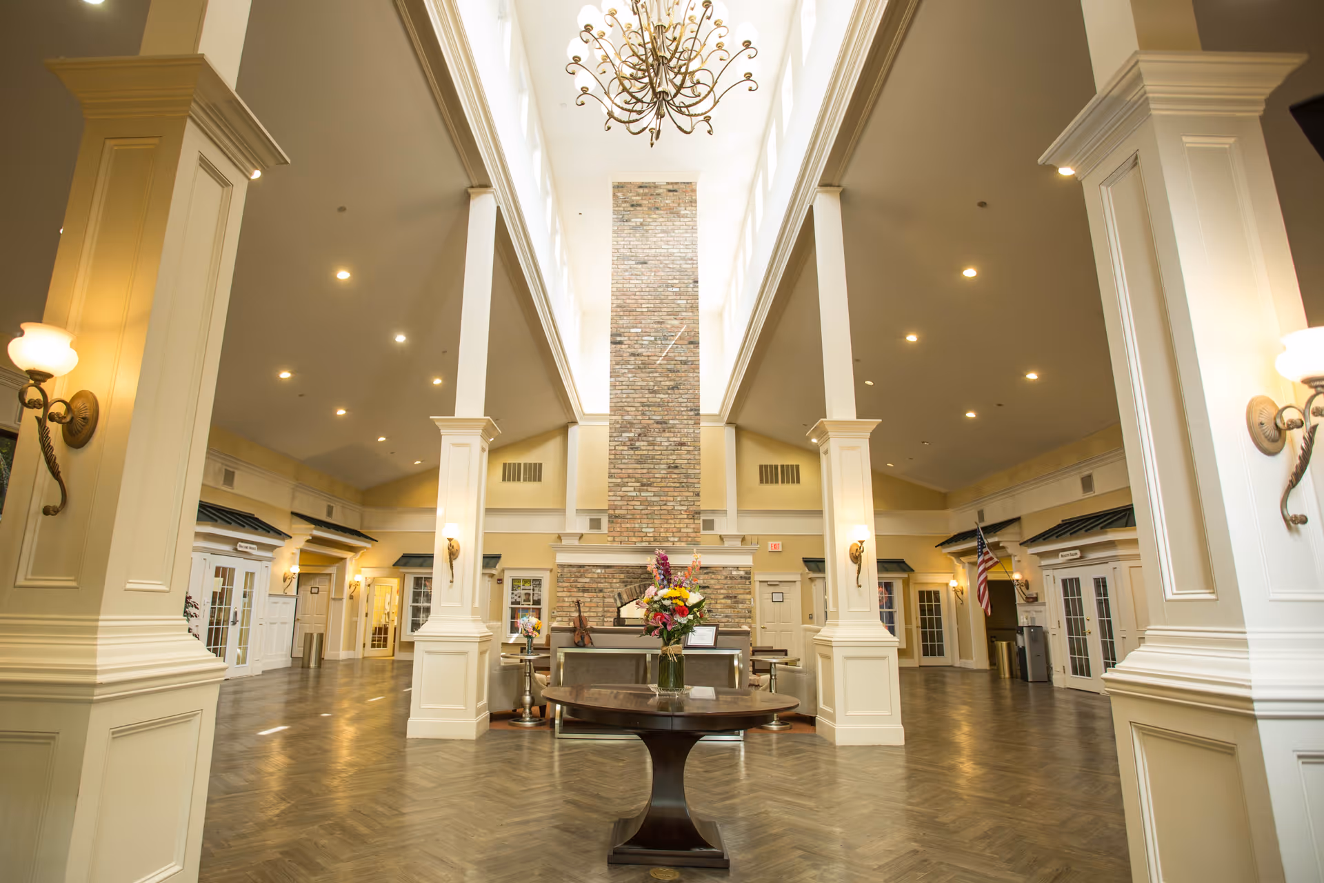 Spacious and elegant interior of a senior living facility with high ceilings, large columns, a central brick fireplace, a chandelier, and a round wooden table with a flower arrangement in the foreground. The room is well-lit with wall sconces and recessed lighting, and there are multiple doorways and seating areas visible along the walls.