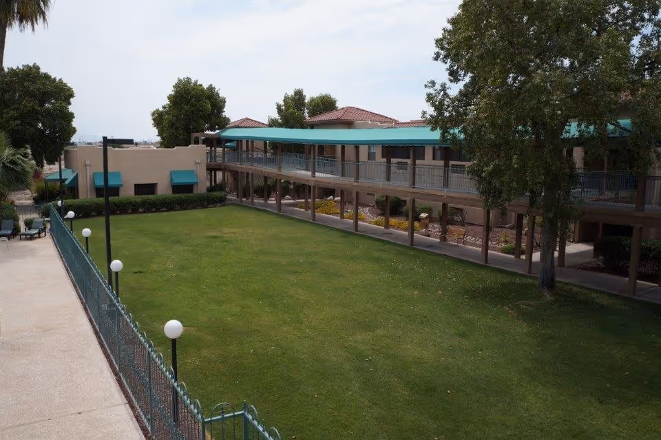 A grassy courtyard bordered by a two-story building with teal awnings, covered walkways, lamp posts and trees.