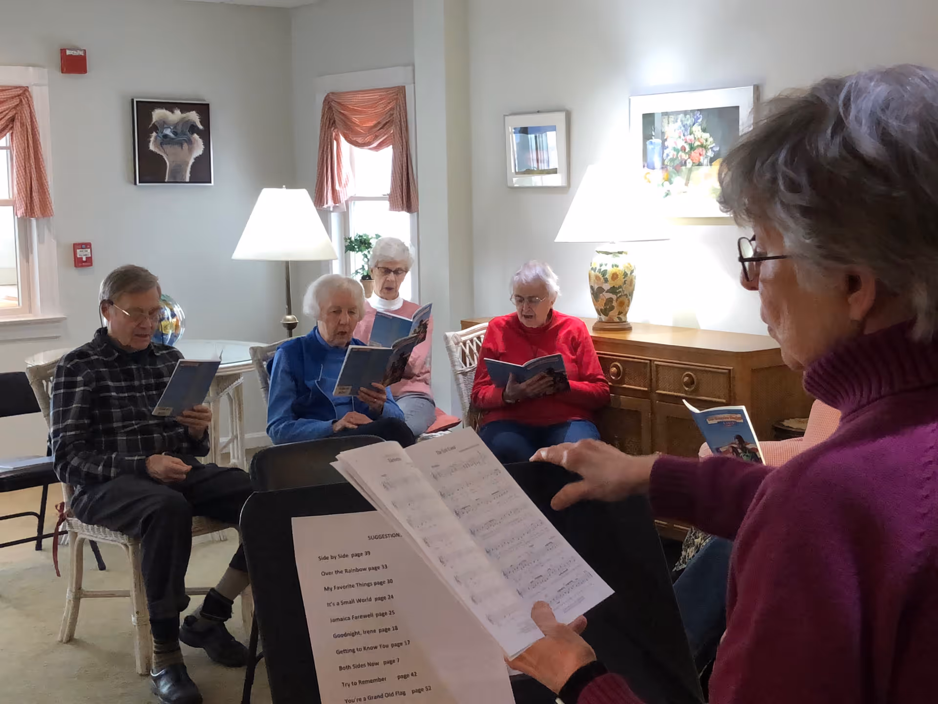 A group of elderly people sitting in a room, holding songbooks and singing together. One person in the foreground is holding sheet music and appears to be leading the group. The room is softly lit with lamps and decorated with framed pictures and curtains.