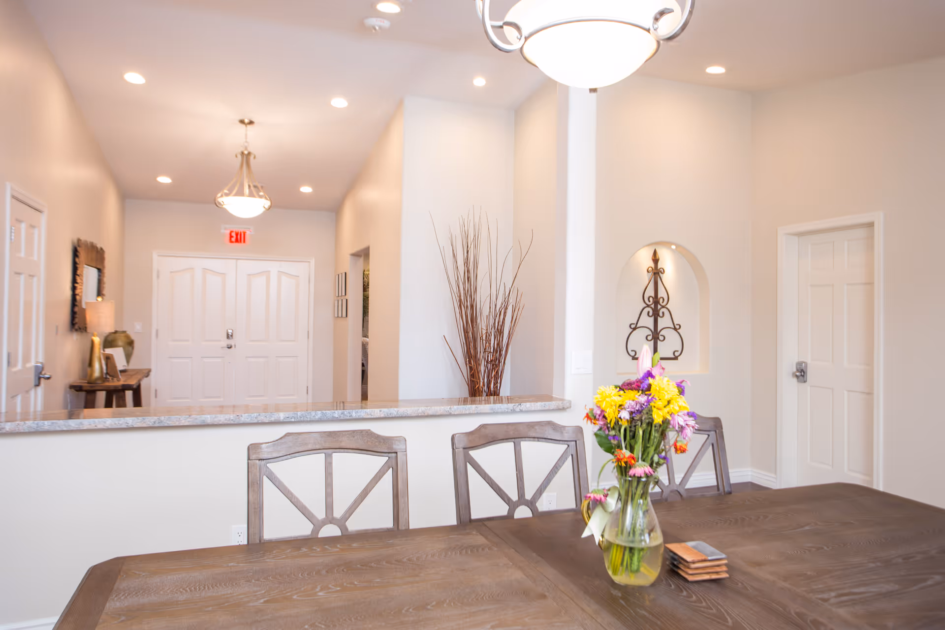Interior view of a well-lit room with a wooden table in the foreground holding a vase of colorful flowers and some coasters. Behind the table are three wooden chairs. The background shows a hallway with white double doors, a side table with a lamp and decorative items, a wall niche with an ornamental iron piece, and a tall vase with dried branches. The walls are painted light beige and the ceiling has recessed lighting and a hanging light fixture.
