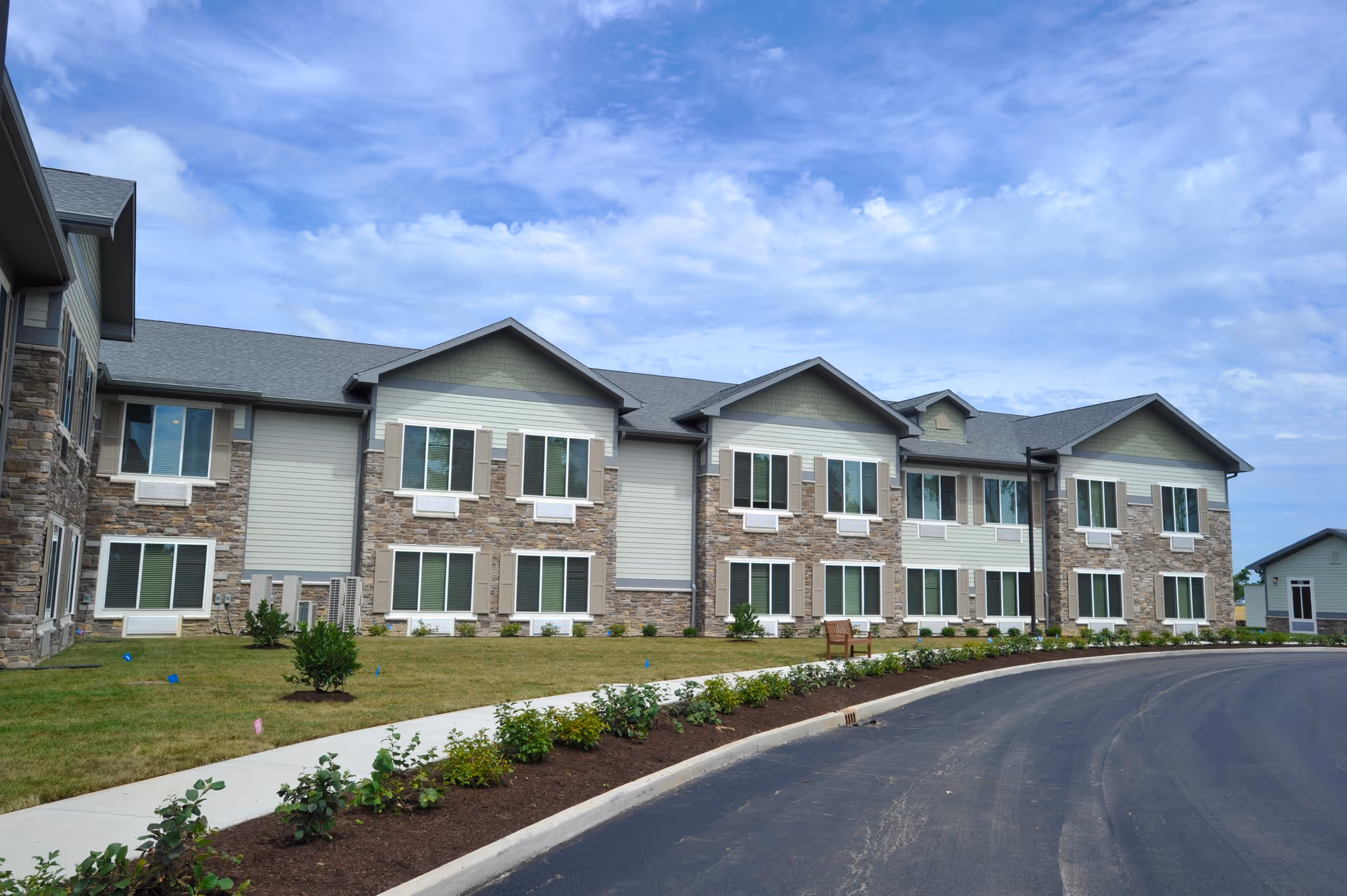 Exterior view of a two-story assisted living facility building with stone and siding facade, multiple windows, a curved driveway, landscaped garden beds, and a partly cloudy sky.