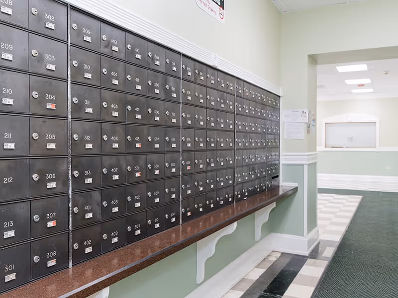 A wall of black metal mailboxes with numbered slots in a hallway of Commonwealth Apartments. The hallway has light green walls with white trim, a brown countertop beneath the mailboxes, and a checkered floor pattern leading to a reception window in the background.
