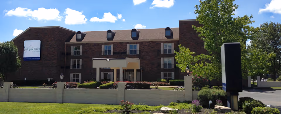 Exterior front view of a three-story brick building with a sign that reads Alpine House of Columbus. The building has multiple windows, a covered entrance, and is surrounded by a low white wall, green grass, bushes, and trees under a blue sky with some clouds.