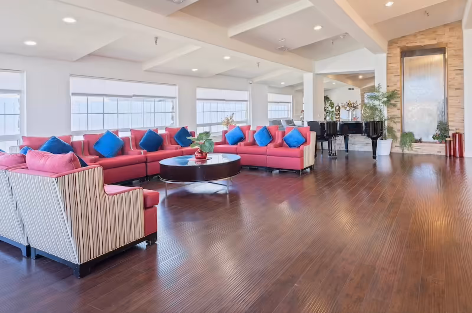 Spacious and bright living room area with large windows, featuring a long red sectional sofa with blue cushions, two striped armchairs, a round dark coffee table with a small plant, a black grand piano, and a tall water feature on a stone wall, all set on a polished wooden floor.