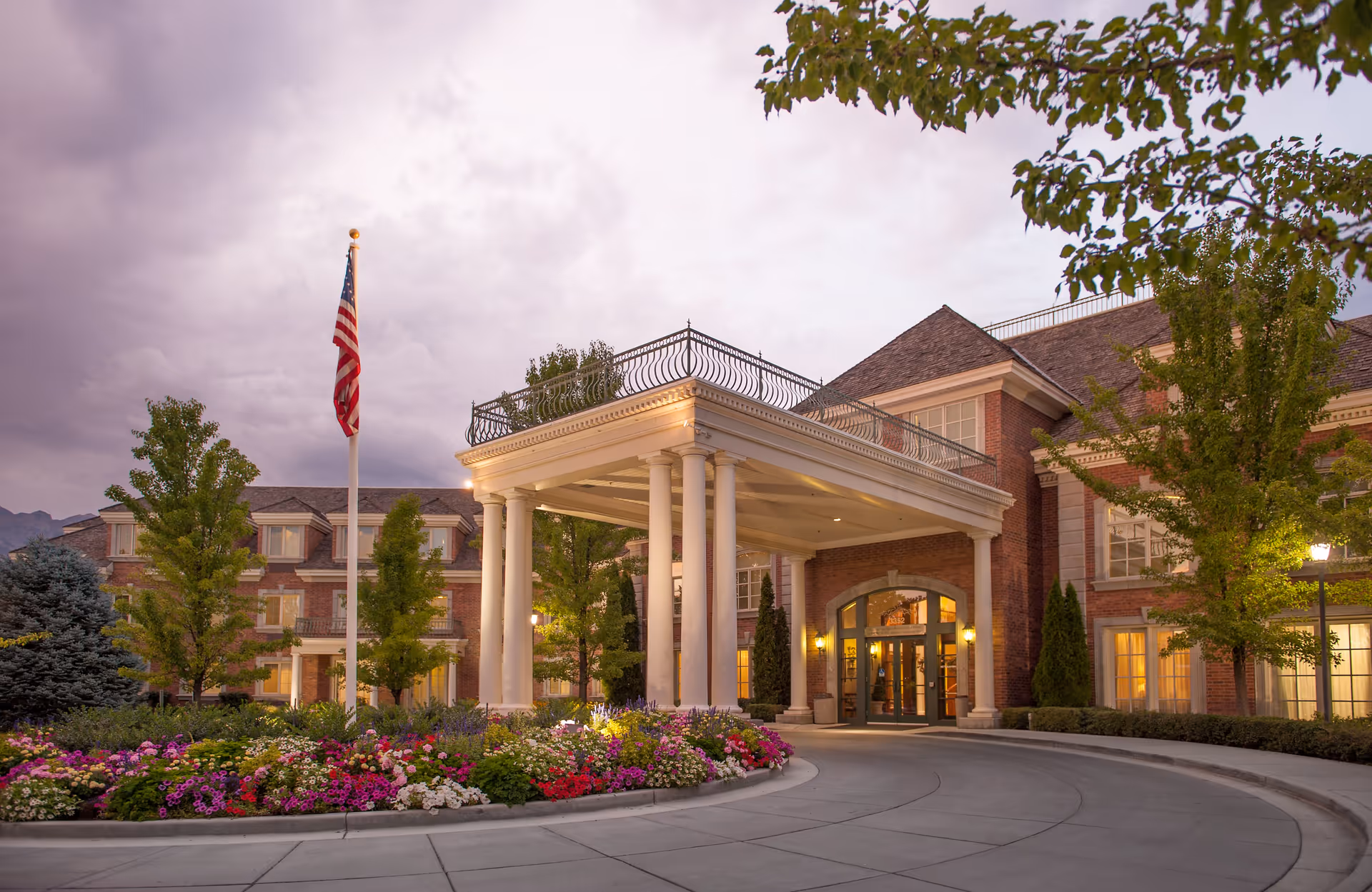 Entrance of a senior living facility named Courtyard at Jamestown during dusk, featuring a large covered driveway supported by white columns, a well-maintained flower bed with colorful flowers, an American flag on a flagpole, and a brick building with lit windows and trees around.