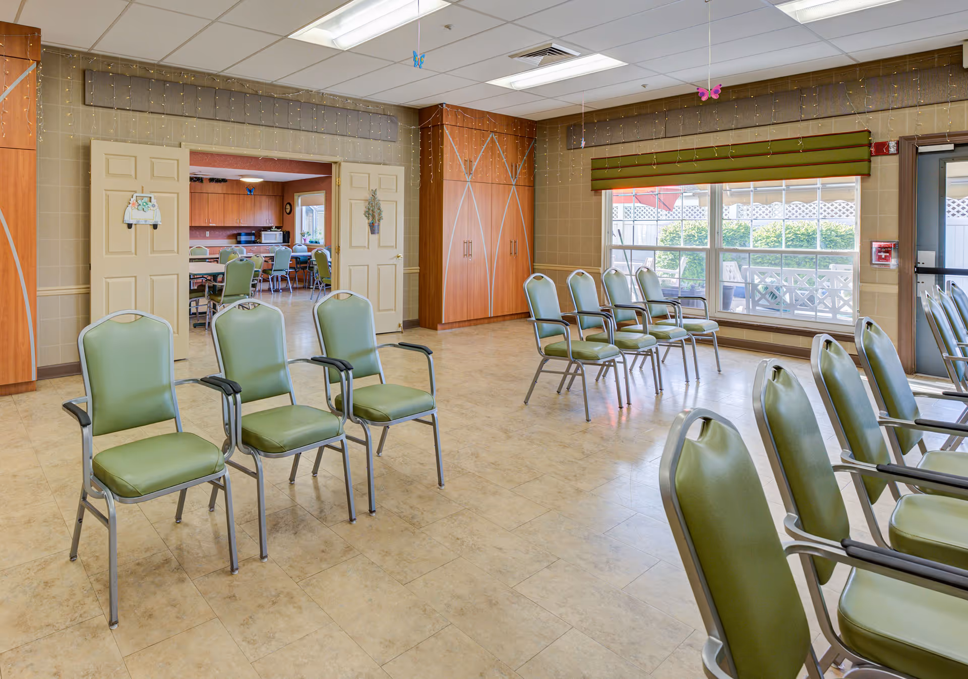 A bright multipurpose interior room with rows of green chairs, large windows, and a doorway opening to a dining/kitchen area.