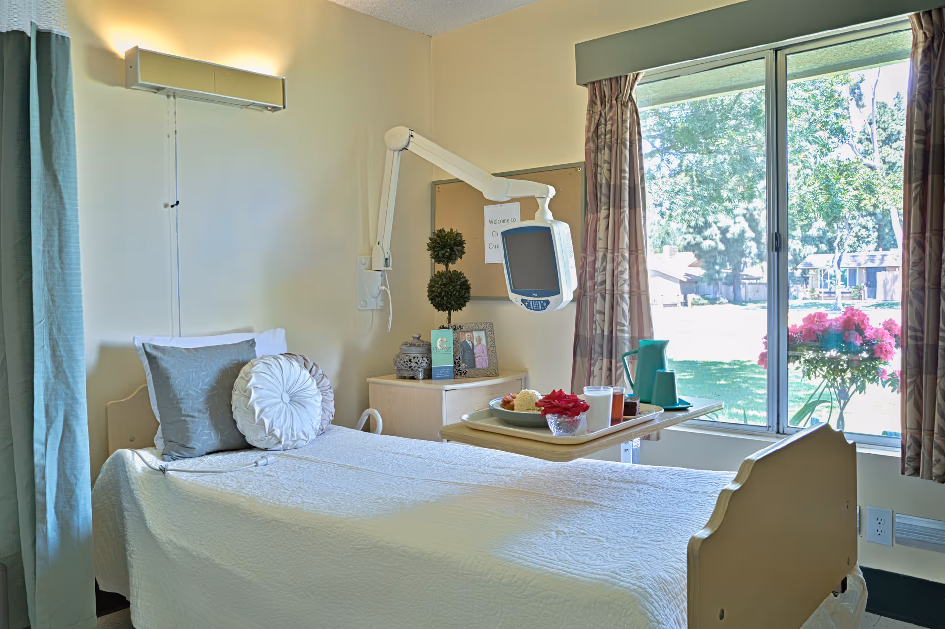 A bright and clean patient room with a single bed covered in white bedding and two decorative pillows. A tray with food and drinks is placed on an adjustable overbed table. There is a wall-mounted medical monitor above a small bedside cabinet with decorative items and a framed photo. Large windows with curtains provide a view of a green outdoor area with trees and flowers.