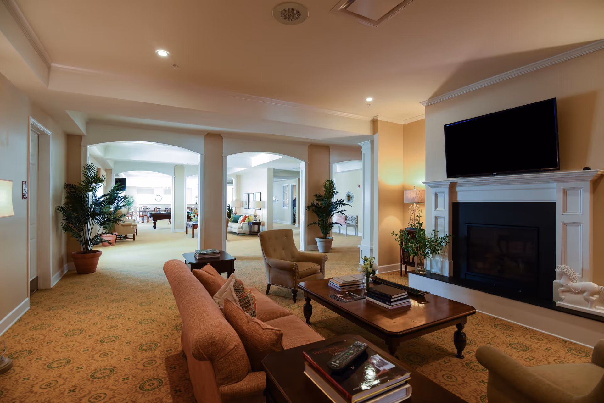A spacious and warmly lit living room area in a senior living facility with comfortable seating including a sofa and armchairs, a wooden coffee table with books and a remote control, a fireplace with a mounted flat-screen TV above it, potted plants, and a carpeted floor. The room extends into another seating area with more chairs and tables visible in the background.