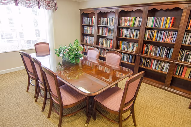 A well-lit room with a large wooden table surrounded by eight upholstered chairs. A floral centerpiece is placed on the table. Behind the table is a large wooden bookshelf filled with books. A window with floral curtains allows natural light to brighten the room.